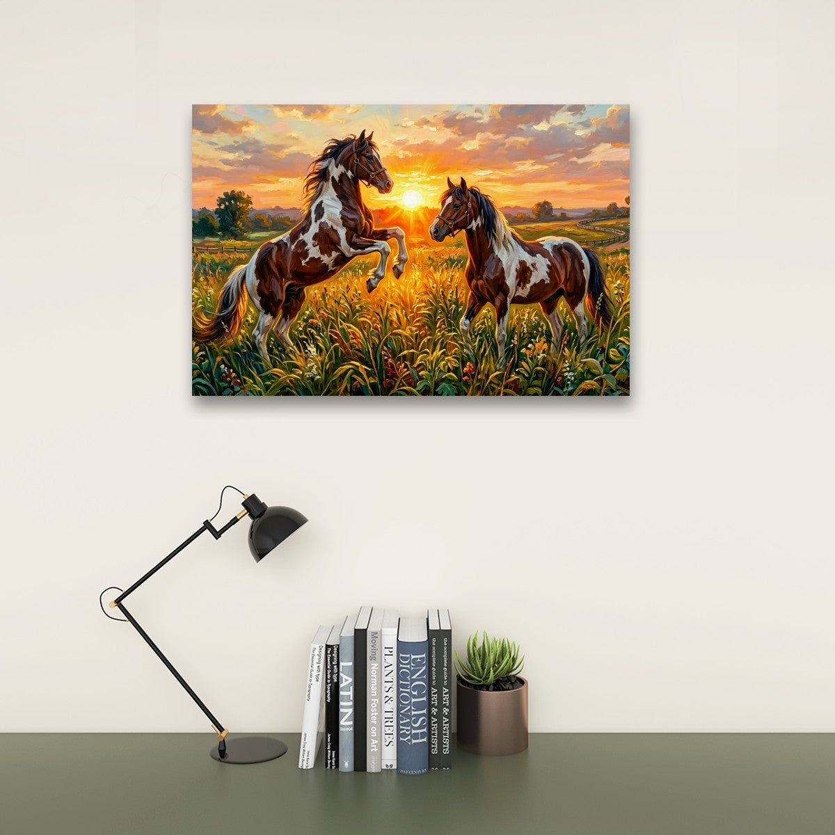 A rectangular wall art print showing two horses in a golden field at sunset, displayed above a desk with a black lamp, books, and a small succulent on a light wall.