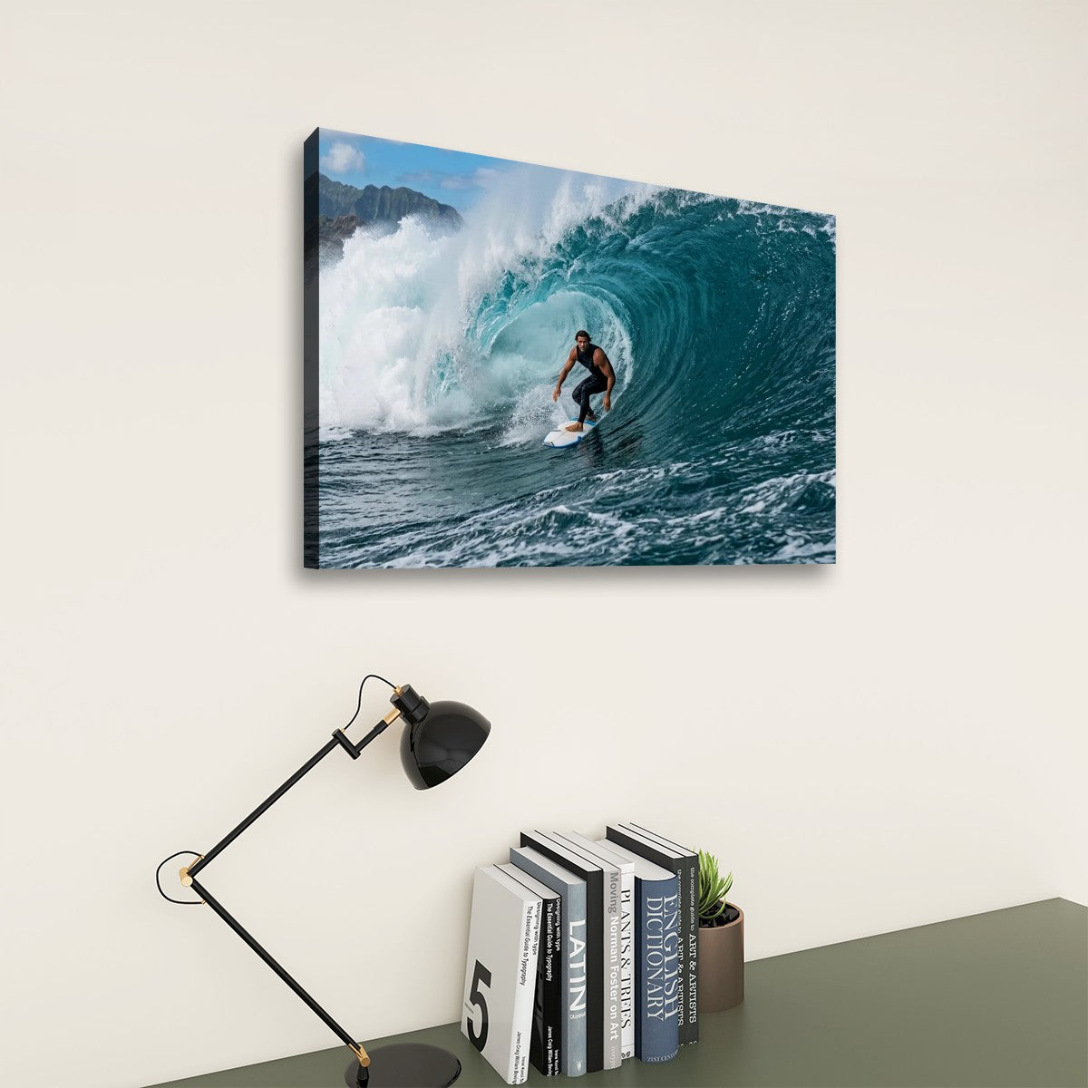 A rectangular wall art print showing a surfer inside a large curling ocean wave, displayed above a desk with books, a small plant, and a black desk lamp against a light beige wall.