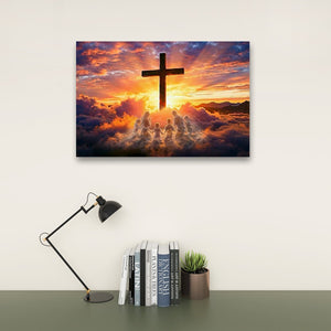 A rectangular wall art print showing a large black cross silhouette against a dramatic sunset sky with clouds and glowing light, displayed above a desk with a black lamp, books, and a small plant.