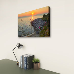 A rectangular wall art print showing a coastal sunset over water, displayed on a white wall above a desk with books, a lamp, and a small plant.