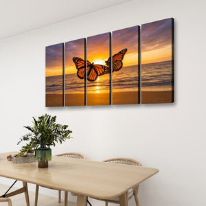 A four-panel canvas wall art set depicting a monarch butterfly over a sunset ocean scene, displayed on a white wall above a light wood dining table with chairs and a green plant.