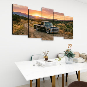 A five-panel canvas wall art set depicting a black pickup truck on a dirt road at sunset, displayed on a white wall above a dining table in a modern room setting.