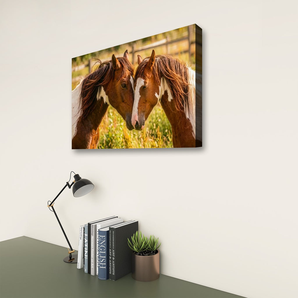 A rectangular wall art print showing two brown horses facing each other in a sunlit field, displayed above a desk with books, a small plant, and a black desk lamp against an off-white wall.