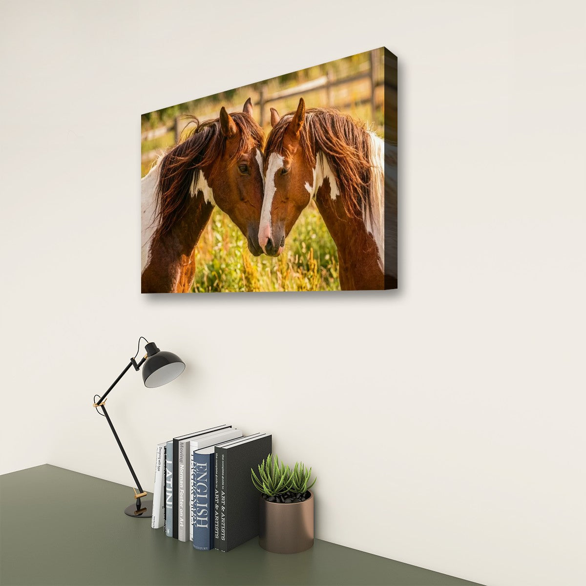 A rectangular wall art print showing two brown horses facing each other in a sunlit field, displayed above a desk with books, a small plant, and a black desk lamp against an off-white wall.