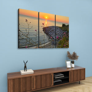 A three-panel canvas wall art set depicting a sunset over a rocky shoreline with flowers in the foreground, displayed on a light blue wall above a wooden console table.