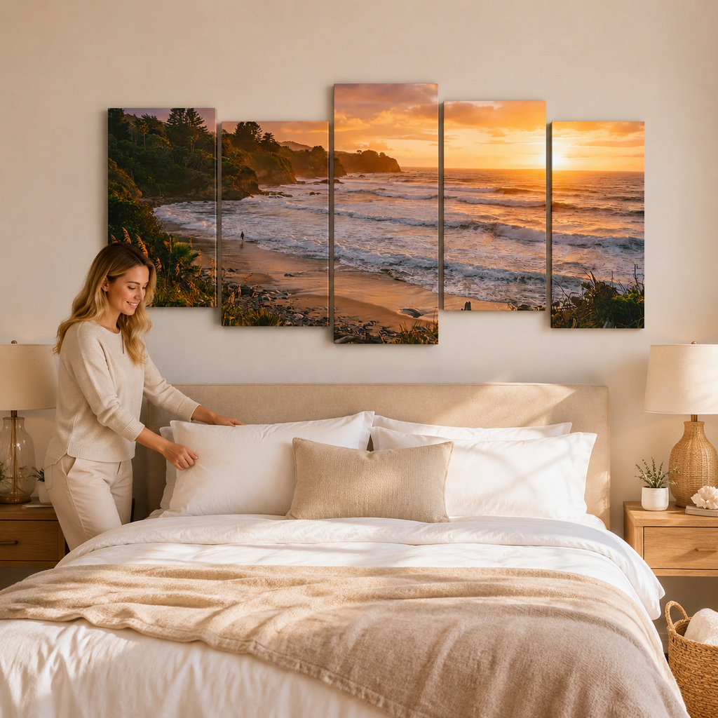 A woman adjusts pillows on a neatly made bed in a warm, modern bedroom. Above the bed hangs a five-panel canvas wall art set depicting a golden beach sunset over the ocean.