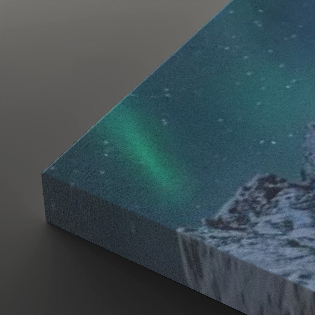 A rectangular canvas or print showing a night landscape with the northern lights in green and teal over a dark blue sky and snowy rocky mountains, photographed at an angle on a dark gray background.