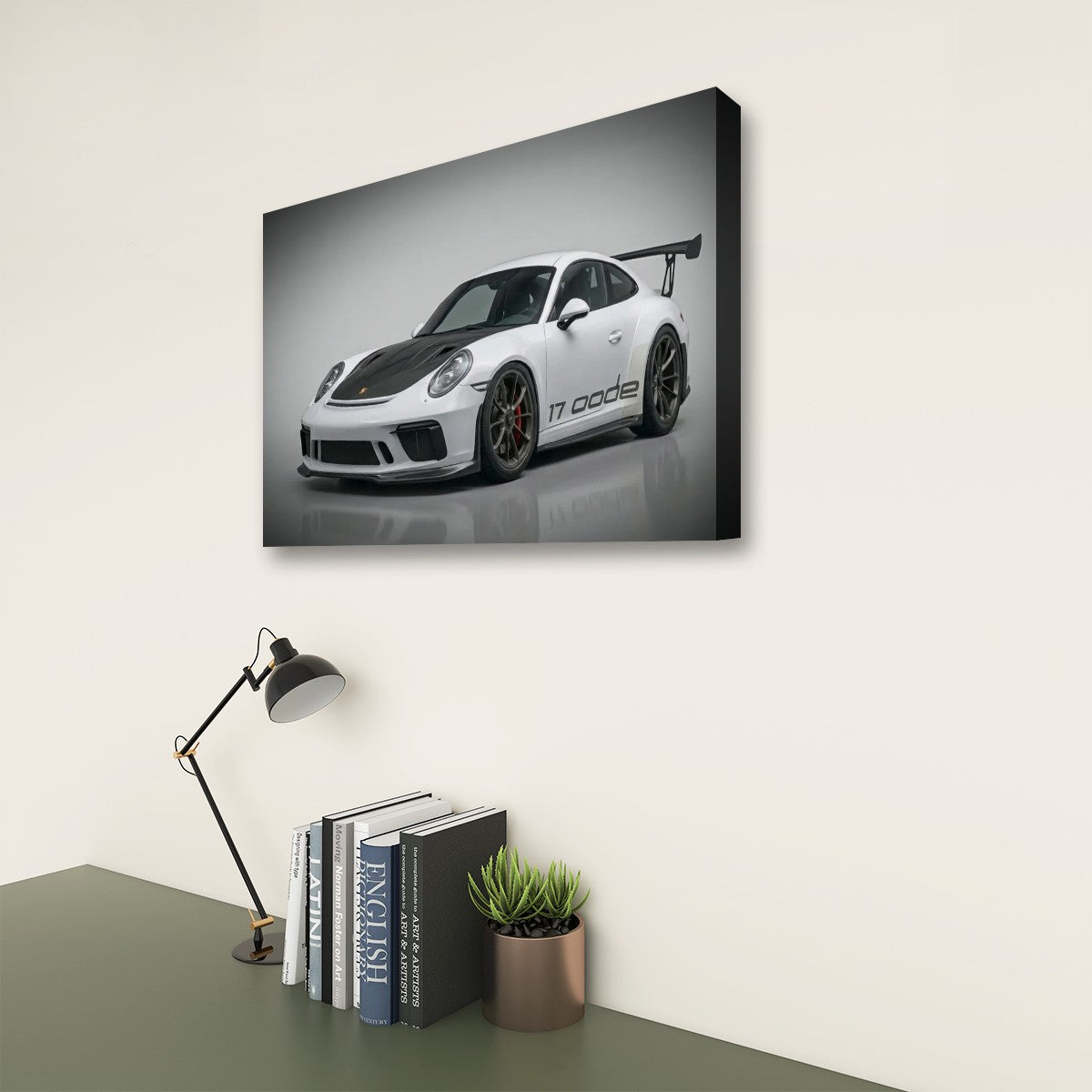 A rectangular wall art print showing a white Porsche 911 Carrera sports car on a gray studio background, displayed above a desk with books, a lamp, and a small plant in a real interior setting.
