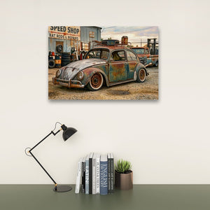 A rectangular wall art print showing a vintage rusted Volkswagen Beetle in a speed shop setting, displayed above a desk with a black lamp, books, and a small potted succulent against a light wall.