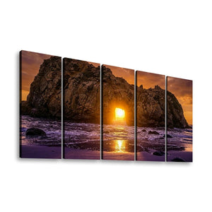 A five-panel canvas wall art set depicting a dramatic rocky coastline at sunset, with the sun low over the ocean and purple-toned waves, shown against a plain white background.