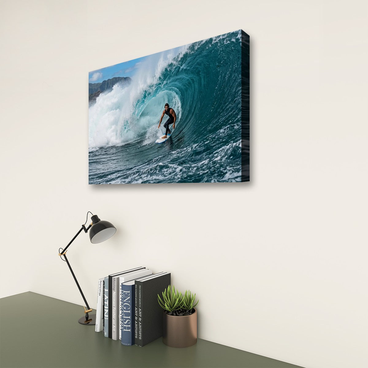 A rectangular wall art print showing a surfer inside a large curling ocean wave, displayed on a light beige wall above a desk with books, a lamp, and a small plant.