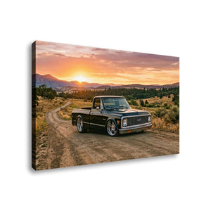 A rectangular wall art canvas showing a black pickup truck parked on a dirt road at sunset, with mountains and trees in the background. The canvas is photographed at an angle against a plain white background.