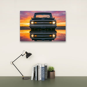 A rectangular wall art print showing the front of a black 1970 Chevy C10 reflected in water at sunset, displayed above a desk with a black lamp, books, and a small succulent in a styled interior setting.