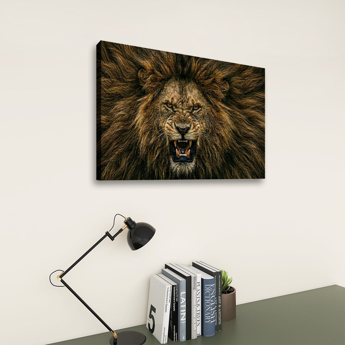 A rectangular wall art print showing a close-up lion face with an open mouth, displayed above a desk with books and a lamp in a styled interior setting.