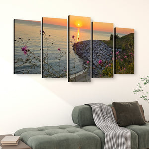 A five-panel canvas wall art set depicting a coastal sunset over the water, displayed above a green sofa in a modern living room setting.
