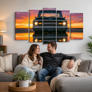 A couple sits on a gray sofa in a living room, smiling and holding mugs, with a large five-panel canvas wall art piece behind them showing a black1970 Chevy C10  pickup truck reflected in water at sunset. The artwork features vivid orange, pink, and purple sky tones.