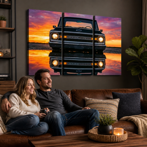 A three-panel canvas wall art print depicting a black vintage 1970 Chevy C10 reflected in water at sunset, displayed on a dark wall above a brown leather sofa with a couple sitting underneath in a cozy living room setting.