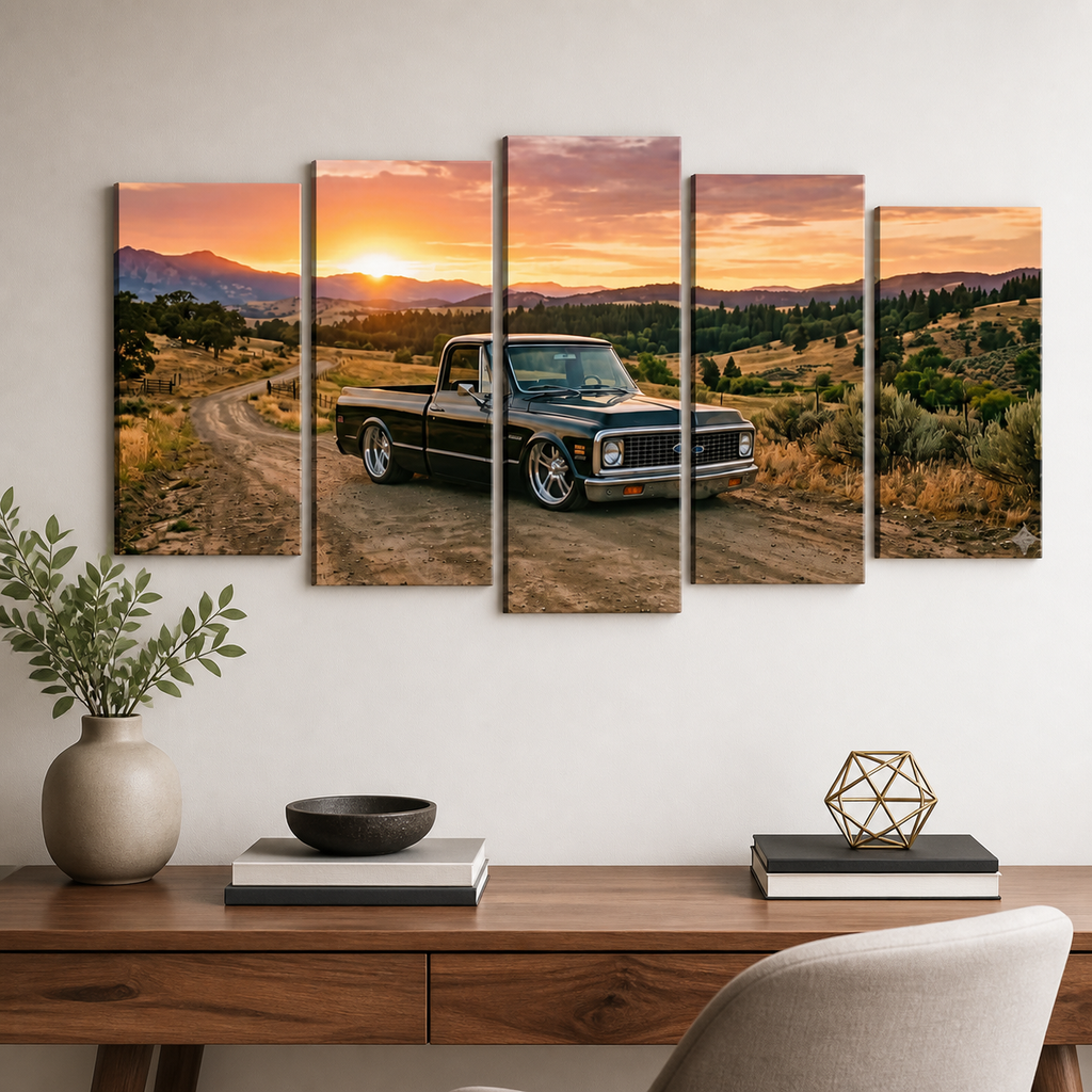 A five-panel canvas wall art set depicting a black vintage pickup truck on a dirt road at sunset, displayed on a white wall above a wooden console table with books, a vase, and a chair.
