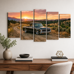A five-panel canvas wall art set depicting a black vintage pickup truck on a dirt road at sunset, displayed on a white wall above a wooden console table with books, a vase, and a chair.