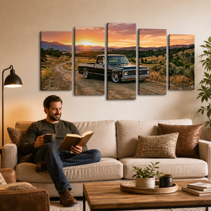 A man sits on a beige sofa in a living room, reading a book and holding a mug. Above him, a five-panel canvas wall art set displays a vintage 1970 Chevy C10 Fleetside pickup truck parked on a dirt road at sunset over a mountain landscape.