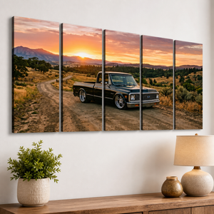 A five-panel canvas wall art set depicting a black vintage 1970 Chevy C10 Fleetside pickup truck on a dirt road at sunset, displayed on a white wall above a wooden console table with a potted plant and a lamp.