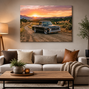 A large rectangular wall art print showing a black pickup truck on a dirt road at sunset, displayed above a beige sofa in a styled living room.