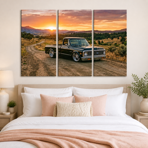 A three-panel canvas wall art print depicting a black vintage pickup truck on a dirt road at sunset, displayed above a neatly made bed in a modern bedroom setting.