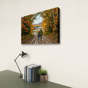 A rectangular wall art print showing a couple walking along a dirt road surrounded by colorful autumn trees, displayed on a light wall above a desk with books, a lamp, and a small plant.