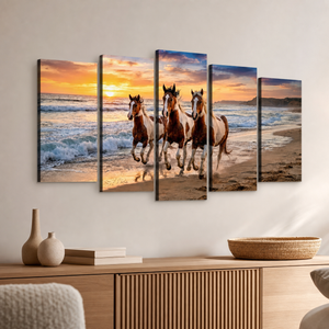 A five-panel canvas wall art set depicting three horses running along a sandy beach at sunset, displayed on a light beige wall above a wooden console table with decorative vases and a woven basket.