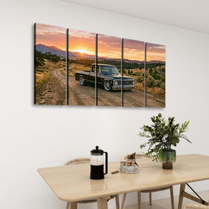 A five-panel canvas wall art set depicting a vintage 1970 Chevy C10 Fleetside pickup truck on a dirt road at sunset, displayed on a white wall above a dining table with chairs, a coffee press, and a plant.