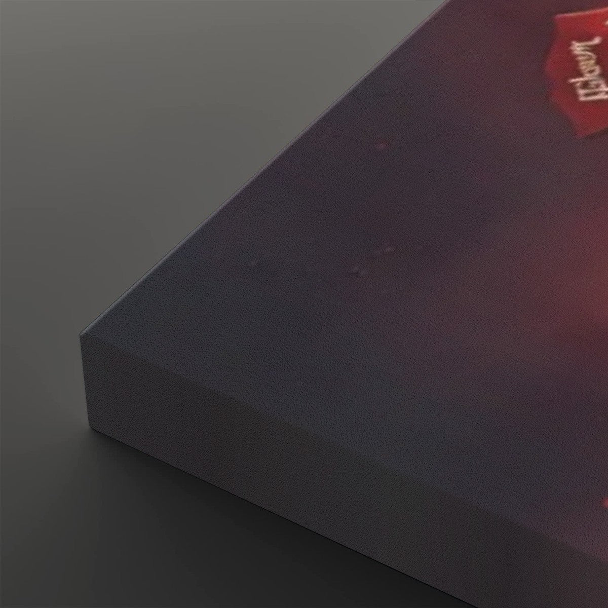 A close-up of a dark rectangular product, likely a book or box, showing a textured dark gray-blue front and a red corner label with white text, photographed on a dark gray background.