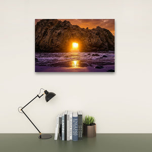 A rectangular wall art print showing a rocky beach at sunset with orange and purple tones, displayed above a desk with a black lamp, books, and a small potted succulent against a light wall.