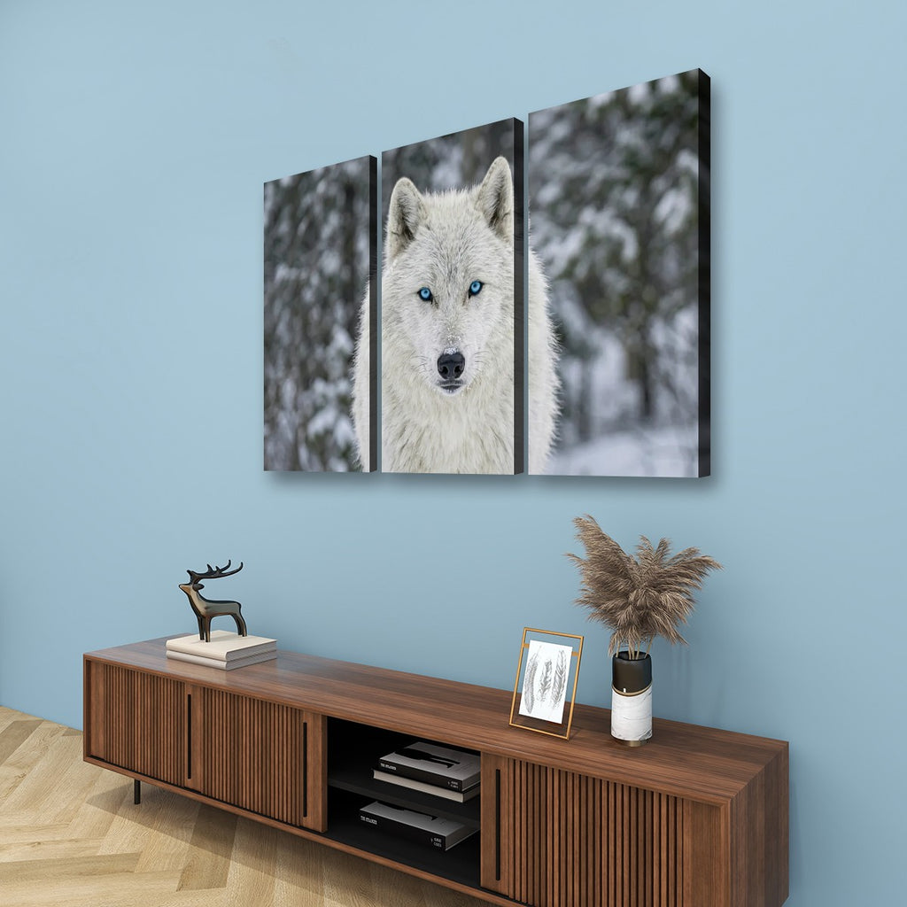 Three-panel canvas wall art depicting a white wolf with blue eyes in a snowy forest, displayed on a light blue wall above a wooden console table.