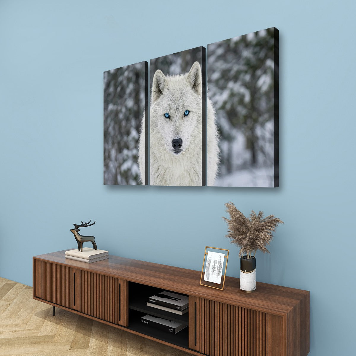 Three-panel canvas wall art depicting a white wolf with blue eyes in a snowy forest, displayed on a light blue wall above a wooden console table.