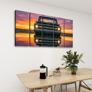 A five-panel canvas wall art set depicting a vintage 1970 Chevy C10 truck at sunset with its reflection on the water, displayed on a white wall above a dining table with a vase and French press.