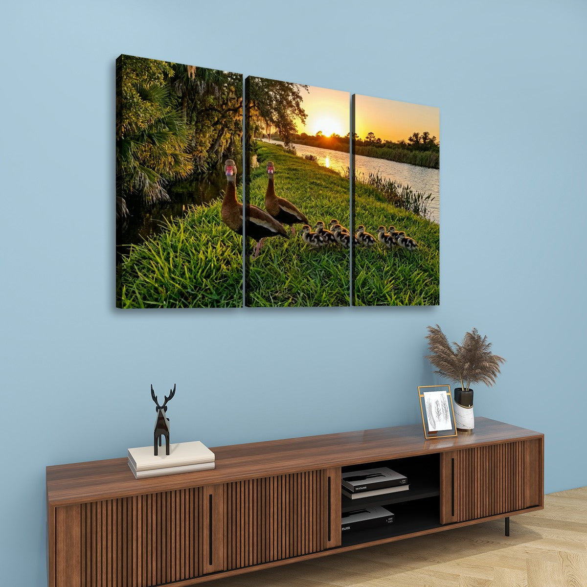 A three-panel canvas wall art set depicting a mother Black-bellied Whistling-Duck and her ducklings at sunset by a lake, displayed on a light blue wall above a wooden console table.