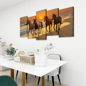 A 4-panel canvas wall art set depicting a sunset beach scene with four horses, displayed on a white wall above a dining table in a modern room setting.