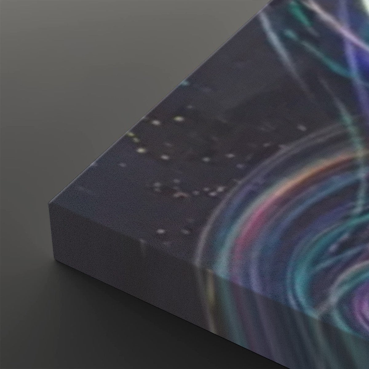 A close-up of a rectangular canvas or print showing an abstract cosmic scene with swirling teal, purple, pink, and blue light trails over a dark starry background, photographed on a dark gray surface.
