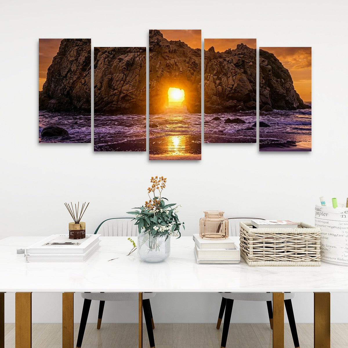 A five-panel canvas wall art set depicting a dramatic rocky coastline at sunset, hung on a white wall above a styled white table with books, a vase, and baskets.