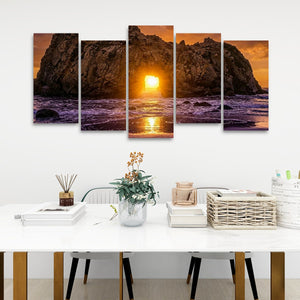 A five-panel canvas wall art set depicting a dramatic rocky coastline at sunset, hung on a white wall above a styled white table with books, a vase, and baskets.