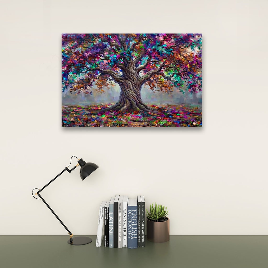 A colorful canvas art print of a tree with textured, painterly foliage in shades of purple, blue, green, orange, and brown, displayed on a light beige wall above a dark green table with a black desk lamp and books.