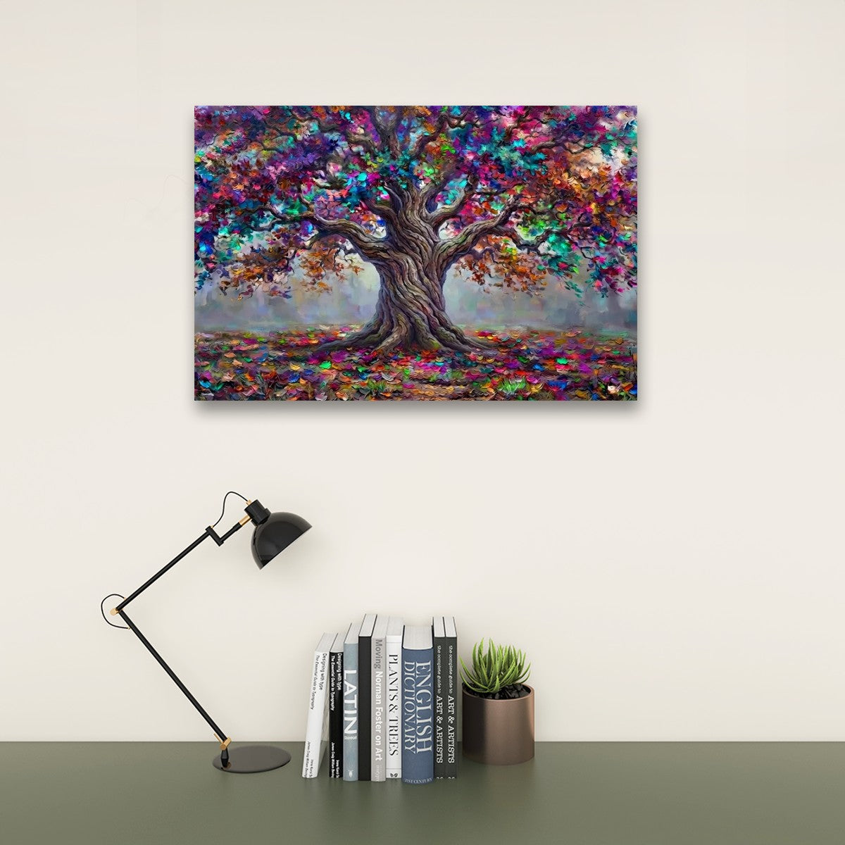 A colorful canvas art print of a tree with textured, painterly foliage in shades of purple, blue, green, orange, and brown, displayed on a light beige wall above a dark green table with a black desk lamp and books.