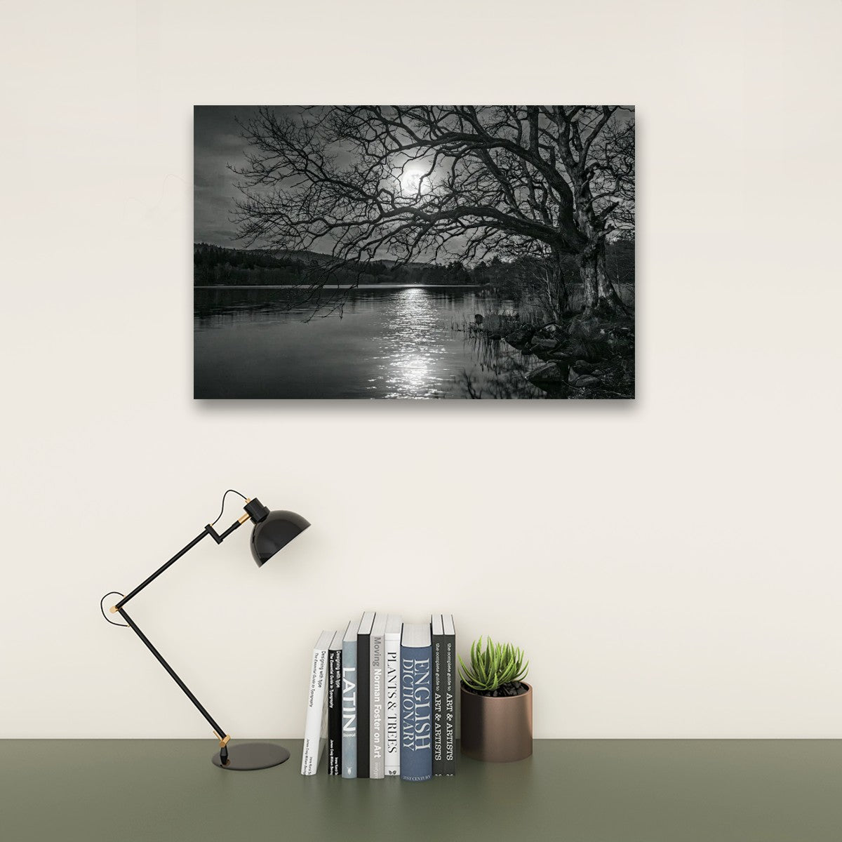 A black and white landscape wall art print depicting a tree by a lake at night under a full moon, displayed on a cream wall above a desk with a lamp, books, and a small plant.