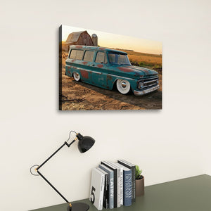 A rectangular wall art print showing a vintage teal 1966 Chevy Suburban Carryall parked in a rural field at sunset, displayed above a desk with books, a small plant, and a black desk lamp against a light wall.