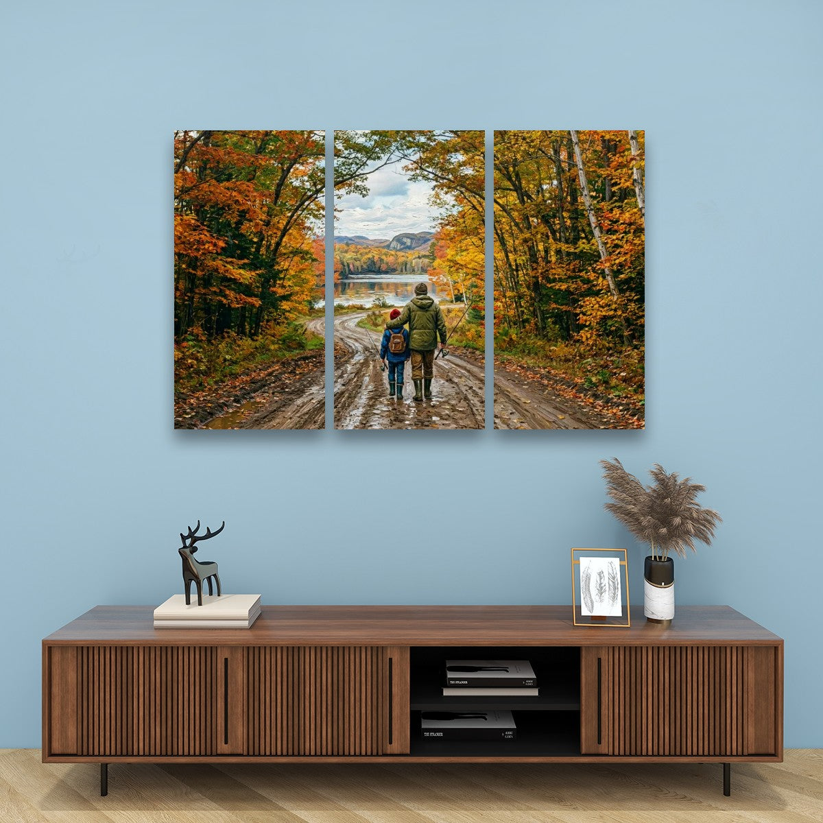A three-panel canvas wall art print depicting a couple walking down a dirt path surrounded by vibrant autumn trees, displayed on a light blue wall above a modern wooden media console.