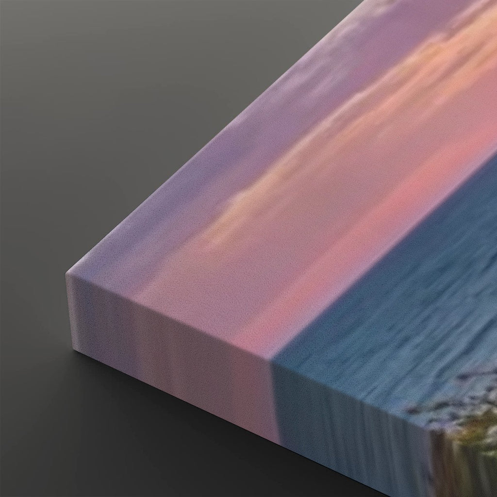A close-up of a rectangular canvas or block showing a scenic sunset landscape with pink and purple sky, blue water, and a small patch of shoreline, photographed on a dark gray background.