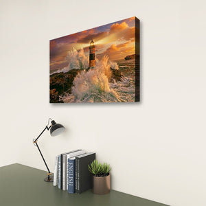 A rectangular wall art canvas showing a dramatic lighthouse scene with crashing waves and a glowing sunset sky, displayed on a light wall above a desk with books, a lamp, and a small plant.