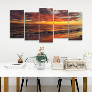 A five-panel canvas wall art set depicting a dramatic sunset over the ocean, displayed on a white wall above a styled dining table with books, a vase, and a basket.