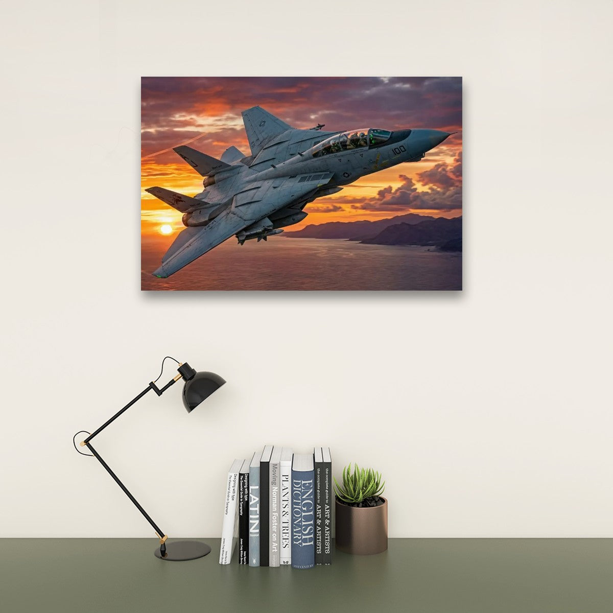 A rectangular wall art print showing a gray military fighter jet flying over water at sunset, displayed on a light wall above a desk with a black lamp, books, and a small plant.