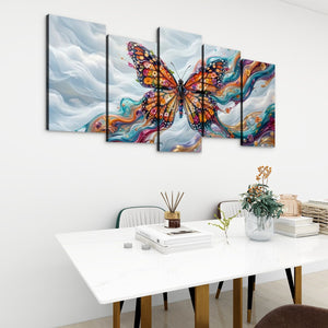 A five-panel canvas wall art set depicting a colorful Monarch butterfly over swirling blue, white, and orange abstract clouds, displayed above a white dining table in a modern room setting.
